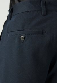 Navy blue trousers made of textured fabric; features a back pocket with a button closure and belt loops along the waistband.