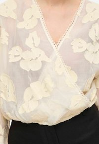Blouse crème transparente avec broderies florales, encolure en V et bordure en dentelle. Tissu léger qui tombe délicatement sur un bas noir ajusté.