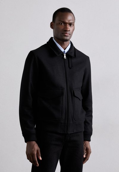 Svart bomberjacka med blixtlås, två framfickor och ribbade ärmslut. Bärs över en blå skjorta med krage.