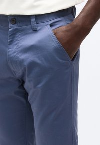 Άτομο με σκουρόχρωμο δέρμα που φοράει μπλε παντελόνι, με το χέρι του τοποθετημένο στην δεξιά τσέπη, πάνω σε λευκό φόντο.