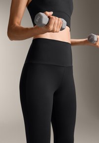 Čierne legíny s vysokým pásom a hladkou, elastickou textúrou, v kombinácii s čiernym športovým podprsenkou. V ruke je držená šedá činká s hmotnosťou 1 kg.