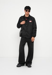 Μαύρο μπουφάν με κόκκινο λογότυπο, μαύρη μπλούζα, φαρδιά μαύρα παντελόνια και μαύρα αθλητικά. Τα αξεσουάρ περιλαμβάνουν γυαλιά ηλίου και τσάντα ώμου.