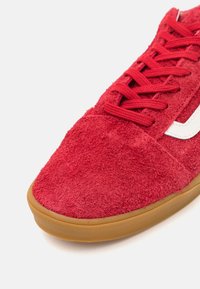 Sneaker in suede rosso con tomaia texture, caratterizzato da una striscia laterale bianca e suola in gomma marrone. I lacci abbinano il colore della scarpa.