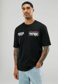 Czarny bawełniany t-shirt z logo "Stranger Things" i "WSQK" w kolorze czerwonym i białym. Luźny krój z krótkimi rękawami, w połączeniu z jasnymi dżinsami.