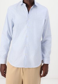 Muž v světle modré a bílé jemně pruhované košili s béžovými kalhotami na jednobarevném bílém pozadí.