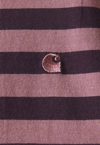 Stribet stof i pink og mørk lilla med et struktureret, broderet logo som accent. Materialet ser blødt og strækbart ud.