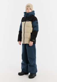 Kurze gepolsterte Jacke mit schwarzen, beigen und taubenblauen Farbbereichen, schwarze Hose und beige Mütze. Mit Reißverschluss und Logo-Details.