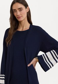 Navy modri cardigan, narejen iz teksturirane tkanine, z širokimi, progastimi rokavi v beli in modri barvi. Odprt spredaj z stranskimi žepe.