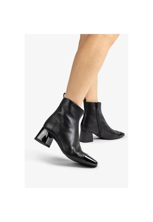Bottines noires à bouts pointus brillants et talons blocs portées sur des jambes croisées sur fond blanc.