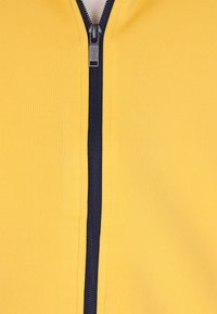 Sárga cipzáros dzseki, texturált anyagból, kontrasztos tengerészkék cipzárral és állgallérral, közeli nézet az elülső részen.