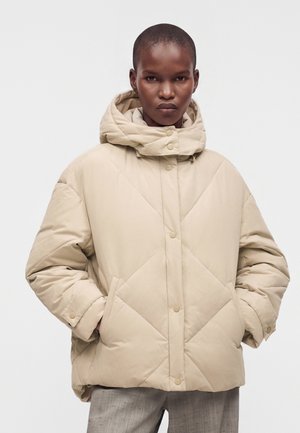 Personne portant une doudoune matelassée beige avec un col montant et les mains dans les poches latérales sur un fond uni.