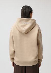 Beige oversized hettegenser med stor hette, ribbestrikkede mansjetter og nederkant, med brodert tekst "LeGer" i hvitt på baksiden. Myk stoff.