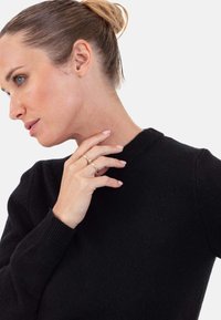 Schwarzer Strickpullover mit rundem Halsausschnitt und gerippten Bündchen. Die Hand des Modells ruht in der Nähe ihres Halses und zeigt zwei Goldringe.