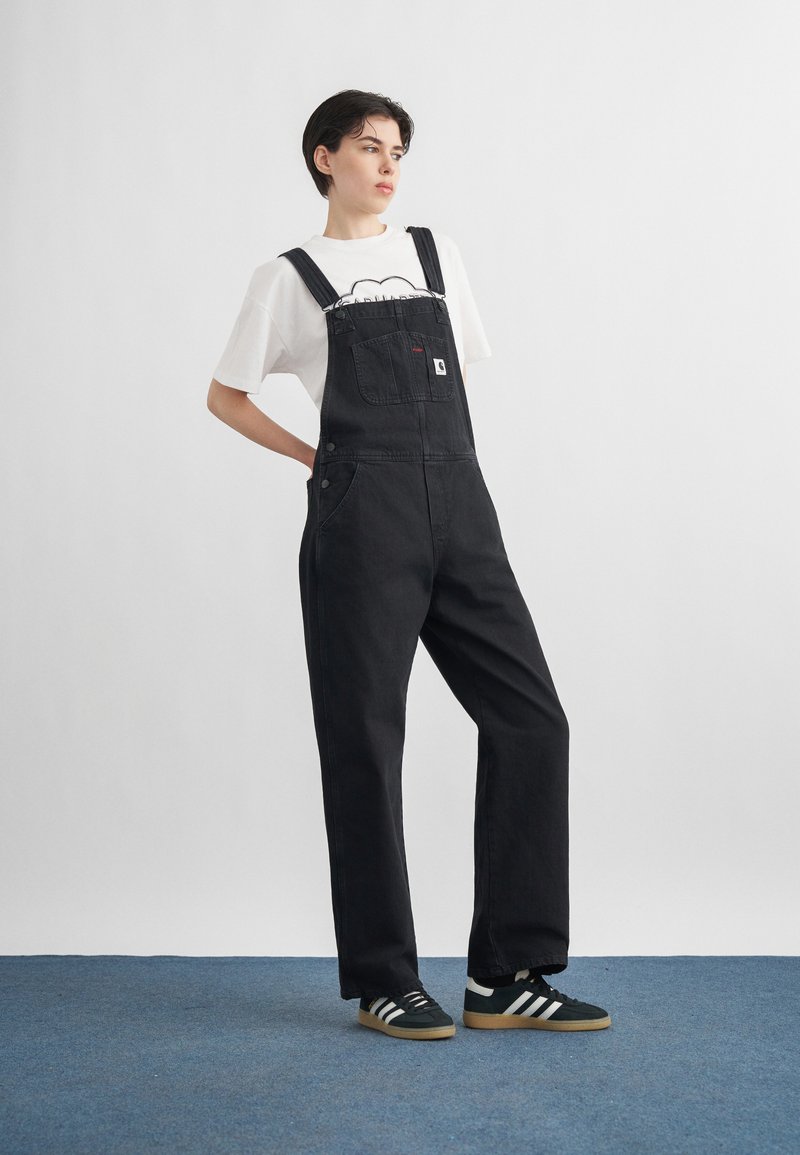 Person mit kurzen dunklen Haaren, die schwarze Jeans-Latzhose, weißes T-Shirt mit Grafik und schwarze Sneakers trägt, steht auf einem blauen Teppich vor einer weißen Wand.