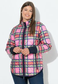 Smilende kvinde iført en farverig pink, grøn og orange ternet quilted jakke med sort lynlås og manchetter, stående mod en hvid væg.