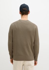 Homme portant un pull côtelé olive et une casquette de baseball marine, vu de dos sur fond blanc uni