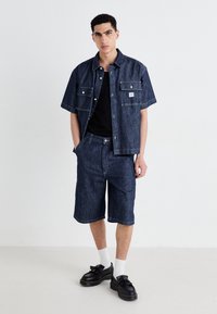 Denim shorts and matching short-sleeve shirt in dark blue, featuring two front pockets and a logo tag. Paired with black shoes and white socks.