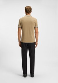 Tanfarbenes Poloshirt mit schwarz-weißer gestreifter Kragen- und Ärmelausschnitt, kombiniert mit schwarzen Hosen und schwarzen Schuhen, von hinten betrachtet.