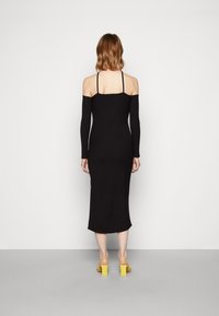 Vestido midi preto com ombros à mostra, mangas longas, silhueta ajustada e textura suave. Combinado com sandálias amarelas de salto bloco.