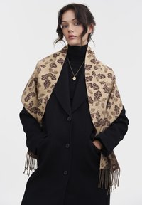 Beige mønstret sjal med brune blade design, med frynset kanter, draperet over en sort frakke. Slim fit, blød tekstur, detaljeret mønster.