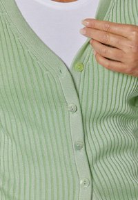 Hellgrüne, gerippte Strickjacke mit V-Ausschnitt, ausgestattet mit sechs passenden Knöpfen und leicht strukturierter Oberfläche. Über einem weißen Oberteil getragen.