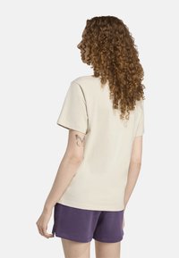 Camiseta de algodón beige con mangas cortas, escote redondo y un corte relajado, combinada con pantalones cortos morados. Textura notable y simplicidad.