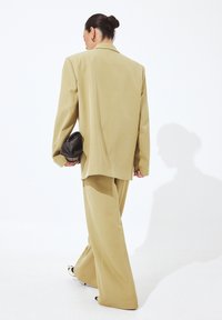 Bežs pārmērīgs uzvalks ar plašām biksēm un saskaņotu žaketi. Strukturēta auduma un minimālisma dizains. Melna soma rokā.