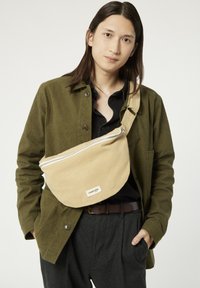 Sac en bandoulière beige avec une fermeture éclair sur le dessus, fabriqué en tissu durable. Présente une petite étiquette de marque et une bandoulière réglable. Porté sur une veste olive.