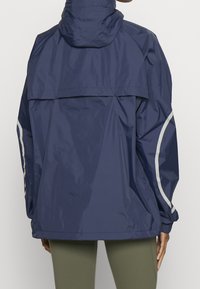 Granatowa wodoodporna kurtka z kapturem, wyposażona w odblaskowe akcenty i luźny krój. Gładka tekstura i casualowy design.