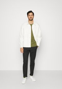 Casaco leve branco por cima de uma t-shirt verde, calças pretas e ténis brancos. As principais características incluem botões, bolsos e punhos enrolados.