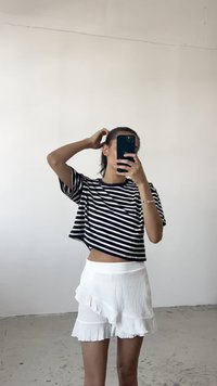 Černobílý pruhovaný crop top s krátkými rukávy, kombinovaný s bílou volánkovou minisukní. Pozadí je jednobarevné a světlé.