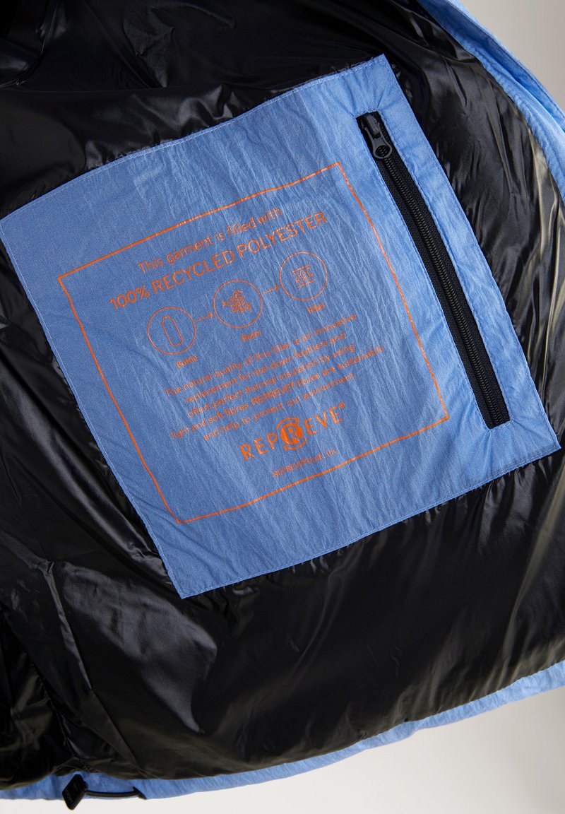 Czarna tkanina wnętrza z jasno-niebieską etykietą z recyklingowanego poliestru. Pomarańczowy tekst zawiera informacje o wypełnieniu i instrukcje pielęgnacji, z zamknięciem na zamek.