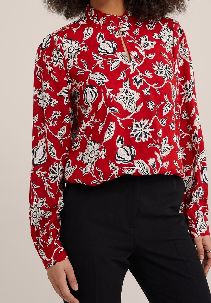 Femme portant un chemisier rouge à manches longues avec des motifs floraux blancs et noirs et un pantalon noir, debout, les bras détendus.