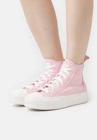 Różowe sneakersy typu high-top wykonane z płótna, z białymi gumowymi podeszwami i sznurowadłami. Posiadają fakturowane wykończenie oraz pętelkę z tyłu.