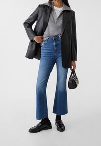 Blazer in pelle nera sopra un maglione grigio, abbinato a jeans a zampa d'elefante in denim blu a vita alta e mocassini neri. Inclusa una piccola borsa nera.