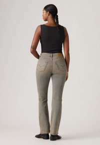 Γυναίκα με πλεξούδες, φοράει μαύρο αμάνικο τοπ, ξεθωριασμένα τζιν με ίσια μπατζάκια, μαύρες μπότες με τακούνια, στέκεται με την πλάτη γυρισμένη σε μονόχρωμο φόντο.