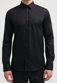 Uomo che indossa una camicia nera a maniche lunghe, aderente, abbottonata, con un colletto classico e pantaloni neri eleganti.
