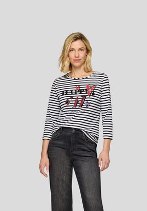 Gestreiftes langärmliges Oberteil in Schwarz und Weiß mit einem grafischen "HAPPY"-Design, das rote und lila Akzente aufweist, kombiniert mit dunkelblauen Jeans.