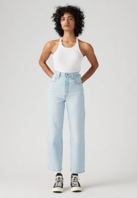 Γυναίκα με σγουρά μαλλιά όρθια, φορώντας λευκό αμάνικο τοπ, ανοιχτό μπλε τζιν ψηλόμεσο και μαύρα ψηλά sneakers με λευκά κορδόνια.