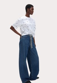 Witte T-shirt met korte mouwen en zwarte bloemmotieven, gecombineerd met ruime blauwe spijkerbroek met trekkoord in de taille. Glad textuur.