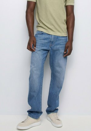 Personne portant un jean bleu clair, une chemise beige à manches courtes et des baskets beiges, debout devant un fond blanc uni.