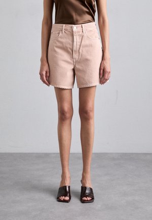 Kobieta w jasnoróżowych, wysokich szortach jeansowych, brązowym topie bez rękawów i ciemnobrązowych butach typu slip-on z odkrytymi palcami, stojąca na szarej podłodze.
