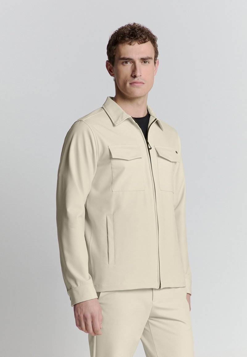 Lichtbeige ritsjack met twee voorzakken op de borst, een overhemdkraag en een slanke, gladde textuur. Gekleed met bijpassende broek.