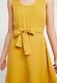 Geel mouwloos jurk met een ronde halslijn en een strikgordel in de taille; soepele, lichte stof; vloeiende A-lijn silhouet.