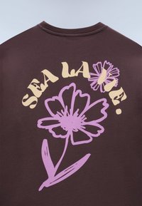 Mørkebrunt skjorte med kremfarget tekst "SEA LA VIE." over to store lavendelfargede blomsterillustrasjoner på baksiden.