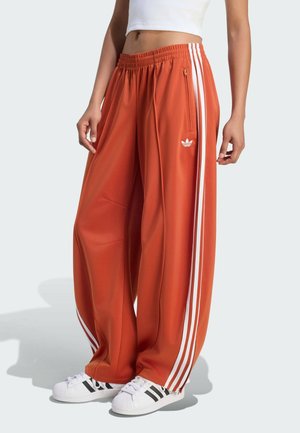 Persona che indossa pantaloni da tuta arancioni larghi con strisce bianche e sneaker bianche con strisce nere, top bianco corto, in piedi su pavimento chiaro.