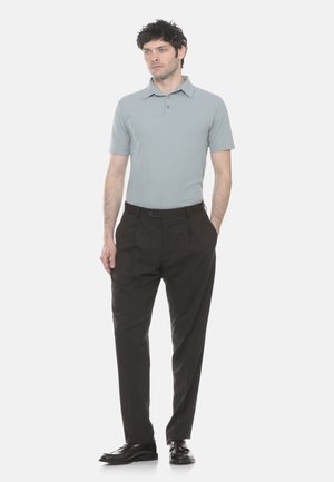 Uomo in piedi con le mani nelle tasche, indossa una polo a maniche corte grigio chiaro, pantaloni marrone scuro e scarpe eleganti nere su sfondo bianco.