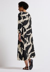 Langes, locker sitzendes Kleid in Beige mit einem auffälligen schwarzen abstrakten Muster. Mit Dreiviertelärmeln und rundem Ausschnitt.