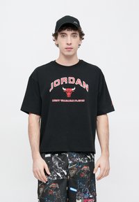 Μαύρο βαμβακερό t-shirt με την επιγραφή "JORDAN" και γραφικό ενός ταύρου σε κόκκινο, άνετη γραμμή, συνδυασμένο με σορτς που έχουν σχέδιο με θέμα το μπάσκετ. Μαύρο καπέλο.