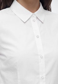 Gros plan d'une personne portant une chemise blanche boutonnée avec un col pointu, montrant la partie supérieure de la poitrine et du cou.